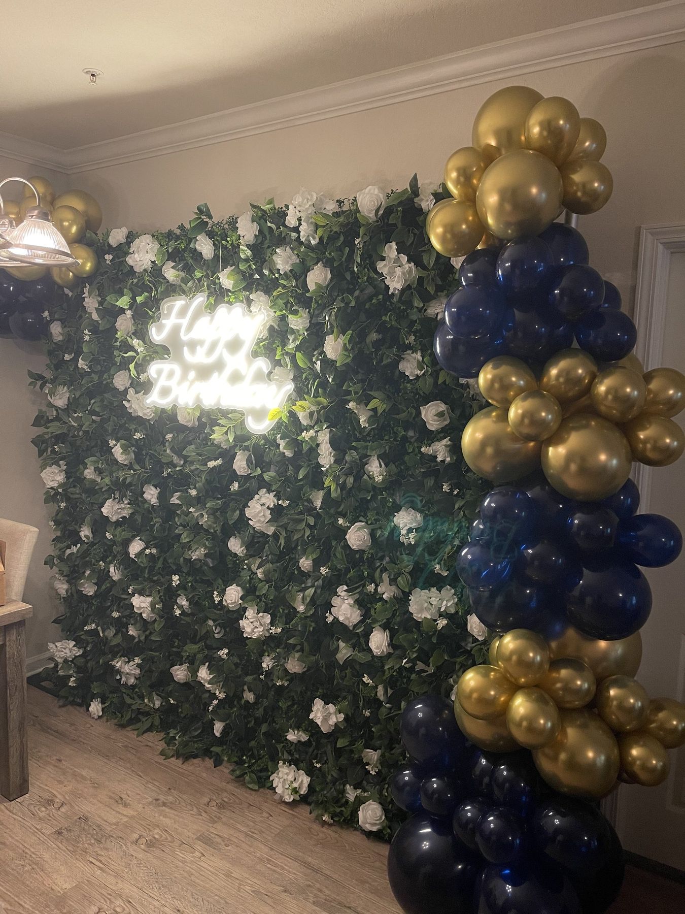 Greenery and white flower wall with a Happy Birthday LED sign and gold and navy balloons at a Tampa party